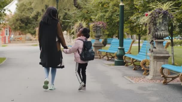 Achteraanzicht Afrikaanse vrouw moeder houdt weinig afro-Amerikaanse meisje met de hand gaan in het stadspark terug naar school studie elementaire klasse les. zwart klein pupil met rugzak gaat naar huis met mama samen — Stockvideo