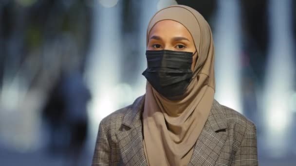 Close-up female face in medical mask in city. Muslim islamic woman in hijab masked lady girl stands evening night urban fountain background outdoors looking at camera posing pandemic coronavirus covid — Stock Video