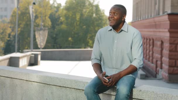 Attractive adult mature African American man sitting resting relaxing outdoors in city on weekend waiting for meeting or date impatiently. Black male cheerfully waving hand welcoming friend colleague — Stock Video