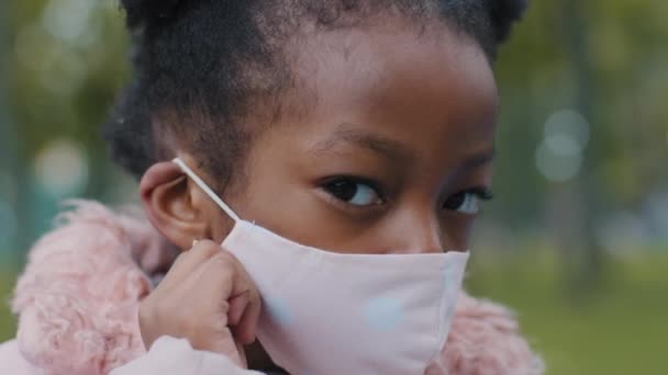 肖像画子供クローズアップ深刻な顔の女の子赤ちゃんアフリカ系アメリカ人の子供少しアフロは、ロックダウンの流行の流行の終了を止めるアレルギーコロナウイルスのカメラの端を見て医療マスクを脱いでcovid — ストック動画