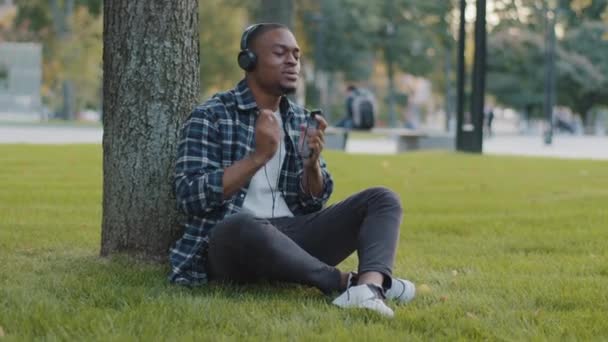 Afro-Amerikaanse man zit in het park op gras in de zomer dag luisteren muziek op koptelefoon zingt lied doen alsof drums spelen met de handen in de lucht. Afrikaanse man student genieten van zingen weekend audio buiten — Stockvideo