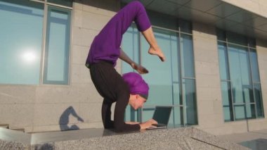Başörtüsü içinde komik bir kız dizüstü bilgisayarında yazı yazan kullanıcı kadın baş aşağı yoga egzersizi yapan asana şehir caddesinde bilgisayarla uzaktan öğrenim görüyor.