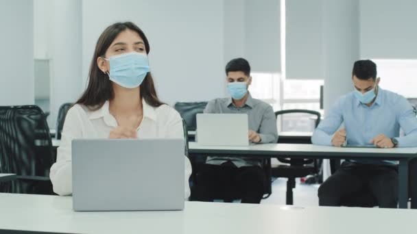 多国籍スタッフは、現代のオフィスでの勤務中にノートパソコンとのコワーキングスペースに座っています。仕事で忙しいお嬢様。顔のマスクを着用し、距離を保ち、流行病の流行による個人の安全 — ストック動画