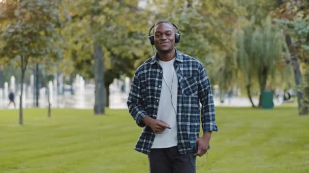 Šťastný aktivní veselý africký Američan kluk student teenager nosí sluchátka chůzi šlapání v parku venku v lese poslech hudby zpěv píseň těší audio. Afro muž ve sluchátkách jde — Stock video