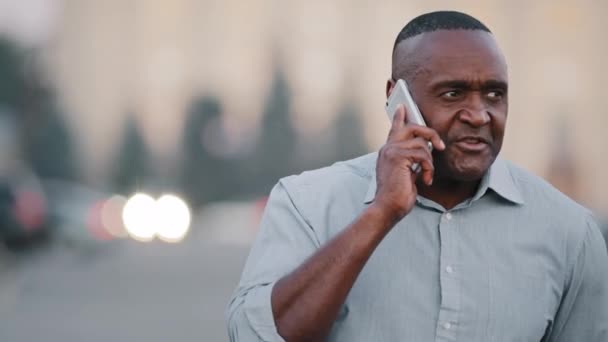 Poważny zaniepokojony dojrzały Afroamerykanin, stojący na zewnątrz, trzymający smartfona w ręku, prowadzi rozmowy biznesowe, negocjuje. Czarny biznesmen używający telefonu komórkowego — Wideo stockowe