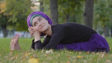 Güzel, genç, İslamcı bir kadın tesettür takıyor. Parkta çimenlerde oturuyor. Göbeğini eğiyor, geriniyor, yoga egzersizi yapıyor.