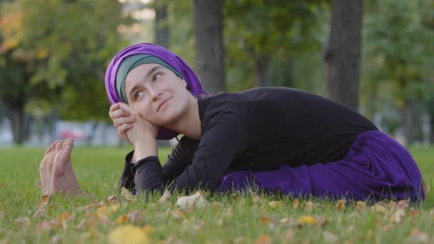Όμορφη νεαρή ισλαμική γυναίκα φοράει χιτζάμπ κάθεται στο γρασίδι στο πάρκο σε εξωτερικούς χώρους κάμψη πάνω στην κοιλιά του σώματος τέντωμα κάνει γιόγκα άσκηση σωματική άσκηση προπόνηση ανάπαυσης χαλαρωτικό χαμόγελο — Αρχείο Βίντεο