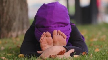 Genç, sakin, tesettürlü, tesettürlü Budist kız parktaki yeşil çimlere oturur. Vücut esneme hareketleri yapar, el ele tutuşur, kameraya gülümser, uluslararası yoga günü egzersizleri yapar.