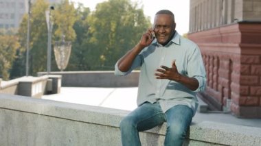 Yetişkin Afro-Amerikan bir adam dışarıda oturmuş cep telefonu tutuyor sinirli sinirli bir şekilde işgüzarla konuşuyor, kötü hava şartları yüzünden üzgün, sözünde durmayan, gecikmiş toplantılar yüzünden üzgün.