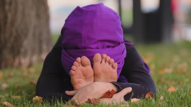 Junge sportlich ruhige, achtsame Buddhistin im Hijab sitzt auf grünem Gras im Park im Freien und macht Stretching-Kurven im Körper streckt Hände zu Füßen und blickt lächelnd in die Kamera, internationales Yoga-Tagestraining — Stockvideo