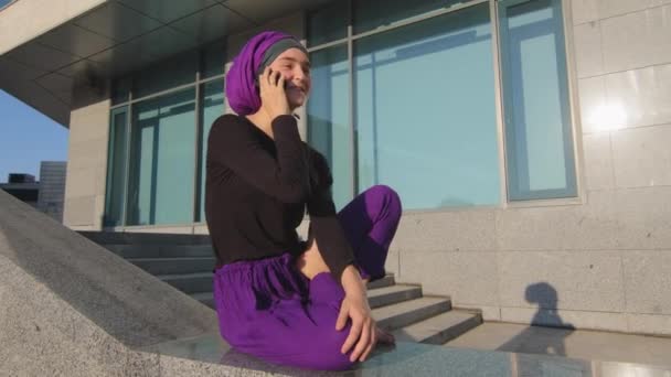 Islámská muslimka studentka dívka v hidžábu sedí na pozadí města budovy venku chatování na mobilním telefonu s úsměvem odpovídání volání mluvit on-line pomocí smartphone mobilní zařízení gadget — Stock video