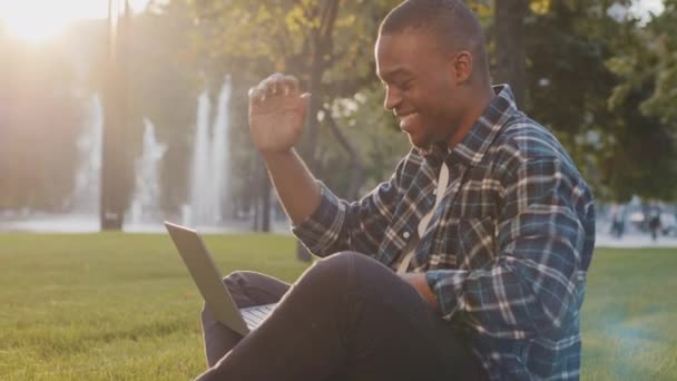 アフリカ系アメリカ人の男が芝生の上の公園に座っていると、ノートパソコンのカメラにこんにちは手を振って話をオンラインビデオ通話は、ネット上の友人とチャットニュースを伝えます。アフリカの男性教師屋外コンピュータとのリモートレッスン — ストック動画