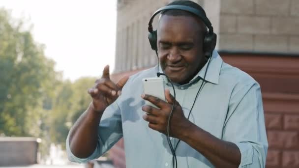 Смешной веселый эмоциональный пожилой афроамериканец в наушниках веселится, слушая музыку с помощью приложения для смартфонов. Активные счастливые пожилые пенсионеры в наушниках танец наслаждаться выходными — стоковое видео