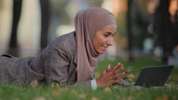 Dost canlısı İslami iş kadını tesettürlü Müslüman kız parkta çimenler üzerinde uzanan bayan öğretmen görüntülü konuşma konferansı yapıyor bilgisayarla uzaktan iletişim kuruyor. — Stok video