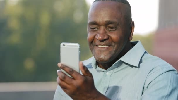 감정적으로 나이든 아프리카 계 미국인 이 스마트폰 카메라를 보면서 핸드폰으로 얘기하는 영상 전화를 하고 있습니다. 행복 한 흑인 남성 중년 은퇴 후 현대 가젯 휴대 전화를 사용하여 야외에서 말하고 있다 — 비디오