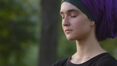 Güzel, genç, İslamcı bir kadın, başörtüsü takıyor açık havada, kadın sakin yüzü kapalı, meditasyon yapıyor derin derin nefes alıyor ve nefes veriyor parkta nefes alma egzersizleri yapıyor meditasyon yapıyor.
