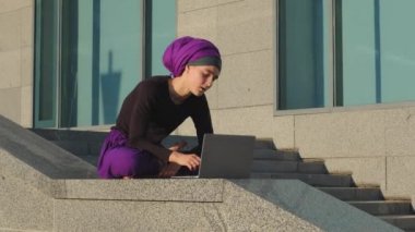 İslami Müslüman iş kadını kullanıcı şehir binasının arka planında oturur dizüstü bilgisayarla çalışır stres yorgunluğu hisseder Lotus pozisyonunda oturur meditasyon yapar