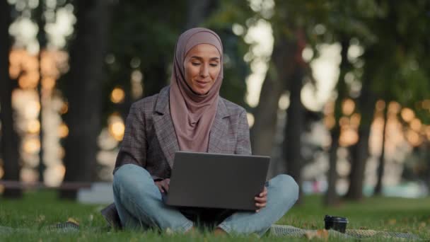 Happy uśmiechnięty przyjazny islamska kobieta muzułmanin dziewczyna w hidżab siedzi na trawie trawnik w parku na świeżym powietrzu w mieście podejmowania rozmowy wideo online czat konferencja rozmowy przez kamerę internetową za pomocą laptopa odległe czat aplikacji — Wideo stockowe