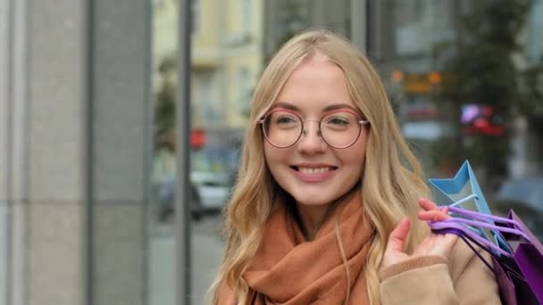 Retrato caucásico feliz mujer hembra shopper sonriendo sosteniendo en mano brillante regalo bolsas joven dama en gafas de pie en la calle y esperando amigo chica disfrutando de descuentos y ventas en el centro de la ciudad — Vídeos de Stock