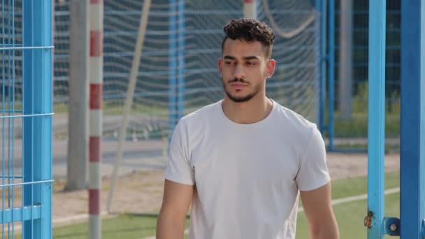 Workout is over. Young guy in sports summer clothes in white t-shirt, middle eastern arab millennial student, indian soccer coach or athlete closing gate of stadium after end of season football game — Stock Video