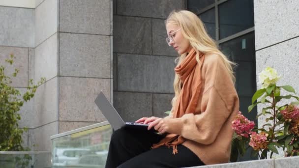 街の建物の近くに屋外に座っている女性のフリーランサーは、ラップトップ上で正常にビジネス女性は、コンピュータを使用して勉強眼鏡をかけて路上でオンラインクライアントとチャット — ストック動画