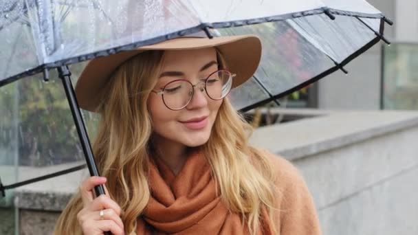 Retrato chica atractiva de pie con paraguas en la ciudad en tiempo lluvioso pensativa joven mujer disfrutando de la lluvia en otoño fuera hermosa hembra caucásica sonriendo posando con sombrero y gafas en la calle — Vídeos de Stock