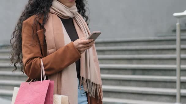 Close-up onherkenbare jonge vrouw shopper in trendy jas en stijlvolle modieuze sjaal wandelen in de stad winkelen met pakketten aankopen met smartphone mobiele telefoon. Korting verkoop zwarte vrijdag — Stockvideo