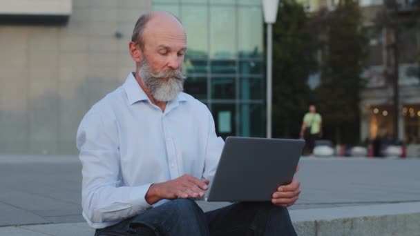 Poważny emeryt rozważać pisarz kreatywny pracownik siedzi na zewnątrz za pomocą laptopa działa zdalnie online. Starszy człowiek dojrzały specjalista z szarą brodą wieku emerytalnego wykorzystuje nowoczesną technologię, aplikacja komputerowa — Wideo stockowe