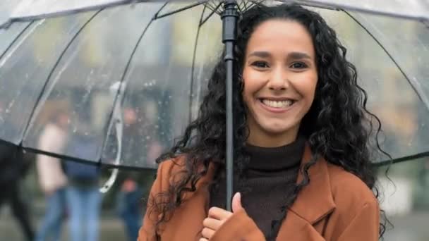 Genç, güzel, kaygısız, mutlu İspanyol kadın portresi. Şemsiyeli, karışık ırk Arap kız yağmurlu havada şehirde duruyor. Sonbaharda yağmurun keyfini çıkarırken sokakta poz veriyor. — Stok video