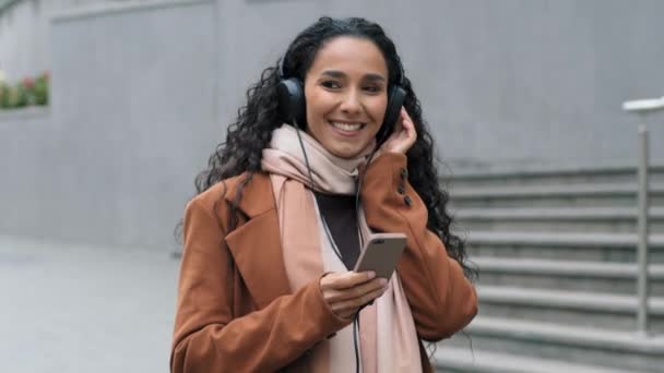 도시 거리를 걷고 있는 아름다운 행복하고 태평 한 처녀 브 런 켓 여성 핸드폰 헤드폰 헤드폰 온라인 오디오 mp3 앱 춤 노래를 즐기는 휴대 전화 이어폰 이어폰을 사용하여 음악을 듣고 있다. — 비디오
