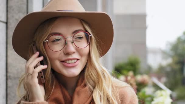 Portrait attractive young girl talking on phone outdoors close-up caucasian happy woman uses smartphone to communicate millennial female with hat and glasses smiling holding gadget makes online order — Stock Video