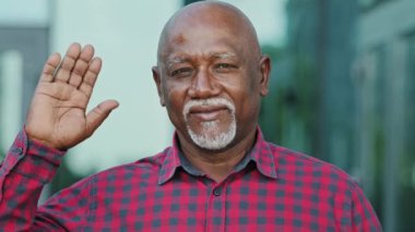 Emeklilik yaşındaki yetişkin, gülümseyen, olgun, dost canlısı kişi avuç içini açıp izleyiciyi selamlıyor. Gri sakallı, yaşlı Afro-Amerikan bir adamın portresi.