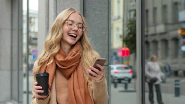Портрет щасливої жінки мобільний телефон на відкритому повітрі несе чашку кави в руці весела дівчина в окулярах, що йде з смартфоном на міському фоні посміхається кавказька леді дивиться мобільний телефон ходьби в місті — стокове відео