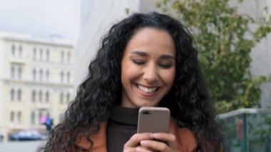 Mutlu Latin Latin kız genç iş kadını şehir caddesinde yürüyor cep telefonu ekranına bakıyor internetten sohbet ediyor sosyal medyada gülen bir mesajla cihaz kullanıyor.
