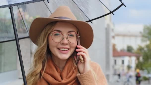 Close-up młoda dziewczyna student spacery w mieście z parasolem kobieta radośnie rozmawiać na zewnątrz telefon komórkowy portret kobieta odpowiedzi połączenie wykorzystuje gadżet pani uśmiechnięta komunikacja na smartfonie na ulicy — Wideo stockowe