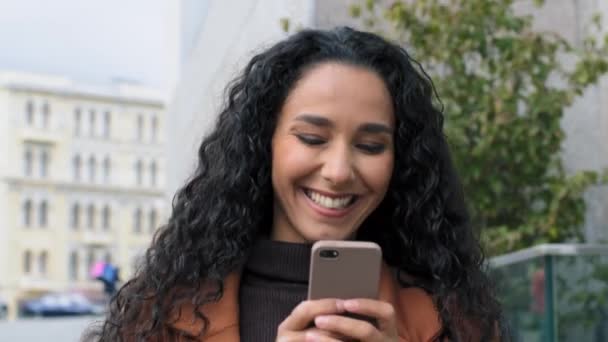 Щаслива латинська іспаномовна дівчина молода бізнес-леді, що ходить вулицею міста, дивлячись на екран мобільного телефону, спілкується онлайн в чистій посмішці, відповідаючи на повідомлення в соціальних мережах за допомогою пристрою гаджета — стокове відео