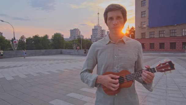 Kharkov, Ucrânia 17.07.2021 Jovem atraente homem de camisa segura ukulele ao pôr do sol ao ar livre. Romântico cara apaixonado se move em direção a câmera, toca instrumento musical popular para namorada no aniversário da data — Vídeo de Stock