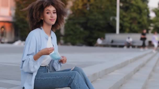 Joven afroamericana chica afro mujer con peinado rizado se sienta en la acera en la ciudad lleva máscara facial médica mirando la cámara muestra gesto bien recomienda usar respirador durante pandemia covid — Vídeo de stock