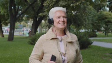 Kulaklıklı, mutlu, kır saçlı, emeklilik yaşında, neşeli bir melodi dinleyen bir kadın. Çekici emekli büyükanne kulaklık takıyor. Parkta rahatlıyor. Modern müzik eşliğinde dans ediyor.