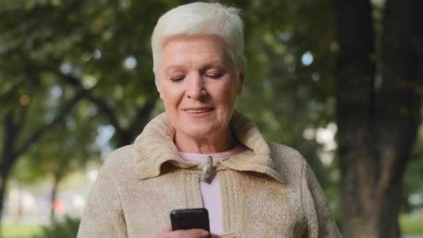 Femme âgée de l'âge de la retraite avec de courts cheveux gris marche dans le parc à l'extérieur tenant le téléphone. Vieille dame élégante souriante regardant des photos à l'écran du smartphone, en utilisant l'enregistrement vidéo et l'envoi de messages — Video