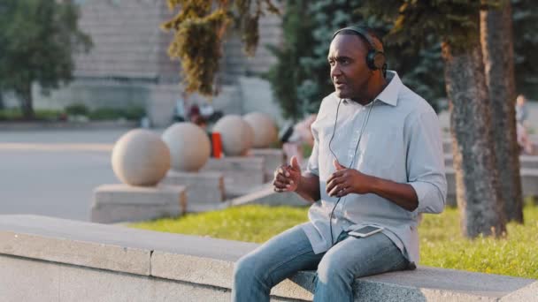 Feliz despreocupado negro emocional senior afroamericano sentado al aire libre escuchando la canción en los auriculares, moviéndose, bailando disfrutando de un gran sonido de los auriculares, utilizando el reproductor de audio de música en el teléfono inteligente — Vídeo de stock