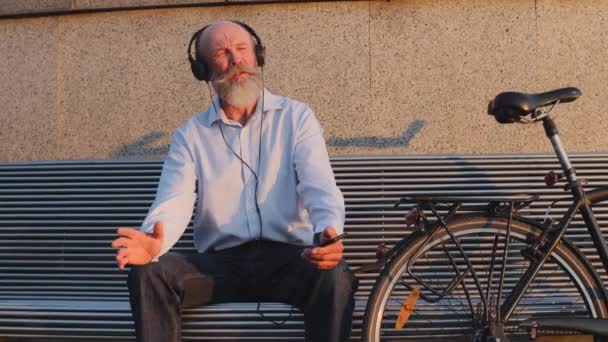 Rolig gråhårig mustasch skäggig cyklist sitter på stadens bänk vilar efter resan. Aktiv gammal man i pensionsåldern, med hörlurar, lyssna glad musik, sjunga njuta av solig helg — Stockvideo