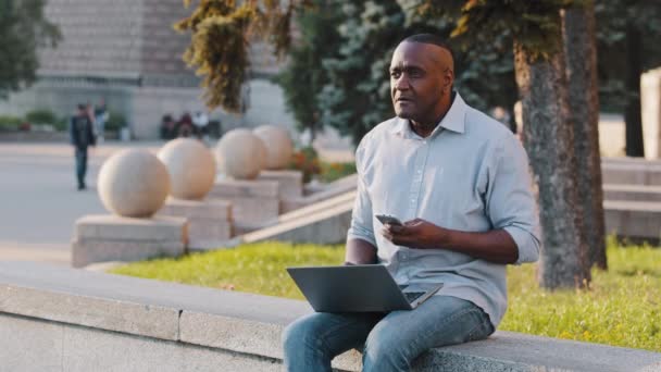 Skupiony starszy czarny biznesmen siedzi na świeżym powietrzu za pomocą laptopa, Afroamerykanin Dojrzałe mężczyzna freelancer odpowiedzi call talk on phone works online Starszy człowiek pracuje na komputerze w miejscu publicznym — Wideo stockowe