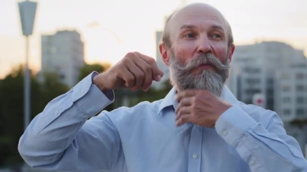 Divertido anciano ríe alegremente se preocupa por peinar bigote gris y barba, visita peluquería y peluquería. Anciano dandy coqueteando mirando a la cámara al aire libre. Puedes verte genial a cualquier edad. — Vídeos de Stock