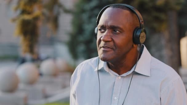 Carefree følelsesmæssige senior afrikansk amerikansk mand sidder udendørs relishing weekend i parken drikker kaffe have det sjovt at lytte sang i hovedtelefoner, bevæger sig, dans nyder øretelefoner med stor lyd – Stock-video