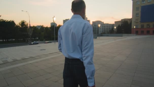 Обратный вид пожилого уставшего кавказца в белой рубашке с деловым портфелем, возвращающимся вечером после тяжелого рабочего дня. Мужчина-пенсионер, пенсионный возраст прогулка на закате на открытом воздухе в городе — стоковое видео