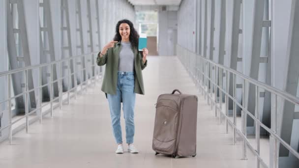 Feliz contento hispano joven viajero turista mujer de pie en el aeropuerto con la maleta muestra apuntando a pasaporte y billete de avión regocija viaje vacaciones en el extranjero demuestra tarjeta de embarque — Vídeos de Stock