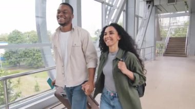 Çok ırklı bir çift Afro adam ve İspanyol bir eş el ele tutuşup bavullarını taşıyorlar havaalanında yürüyorlar mutlu bir şekilde gülümsüyorlar mutlu bir yolculukla zafer jestleri yapıp balayını kutluyorlar.
