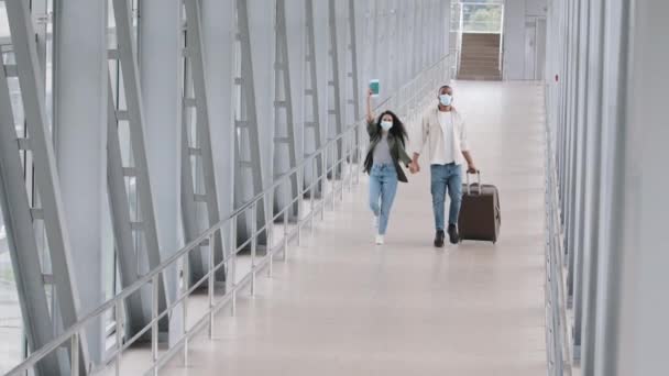 Multi-etnisch paar pasgetrouwden Afrikaans man en zijn paniekerige vrouw gelukkige echtgenoten dragen gezicht beschermende medische maskers lopen springen in terminal van de luchthaven dragen koffer genieten reizen huwelijksreis — Stockvideo