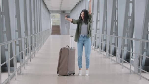 ハッピーガールの女性の乗客は手でスーツケース保持飛行機のチケットで空港に立つオンラインビデオブログvlog会議コールジャンプ携帯電話のカメラにパスポートを示す旅行を喜ぶ旅行 — ストック動画